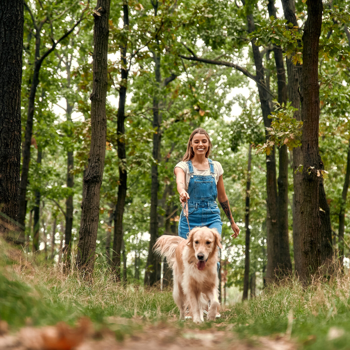 A cheerful and lively young woman is joyfully walking her golden retriever through a vibrant, lush green forest, fully immersed in the beauty and tranquility that nature has to offer at this moment