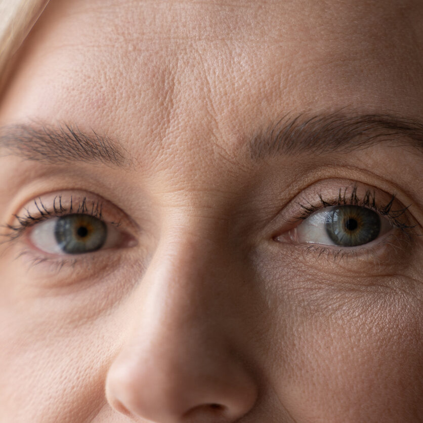 A close-up view captures the top half of a womans face, revealing her striking blue eyes, soft blond eyebrows, and delicate skin texture. The focus on her eyes creates an intimate and introspective mood.