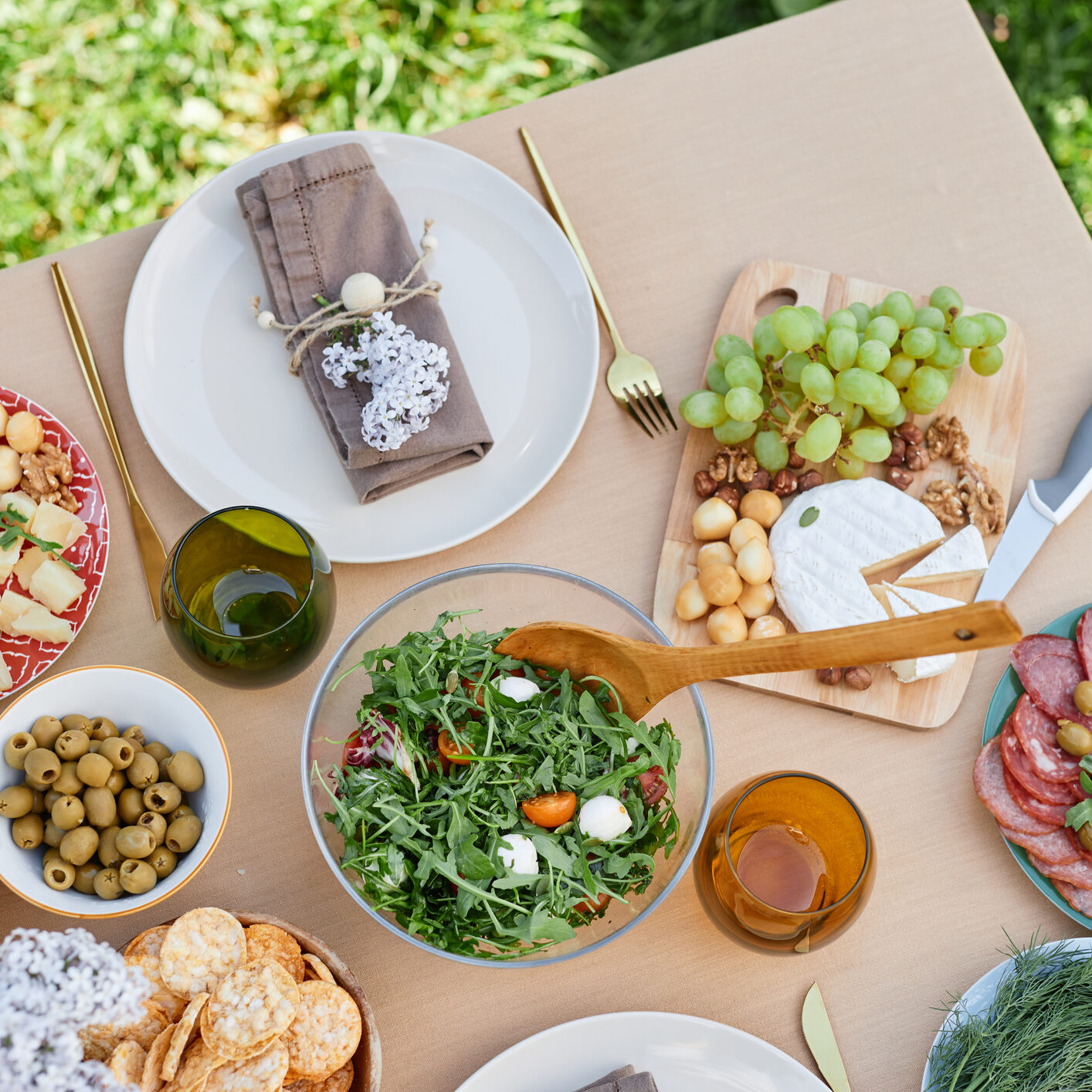 Scenic outdoor picnic setting featuring variety of appetizers including cheese, olives, and fresh salad placed on wooden table without using natural light